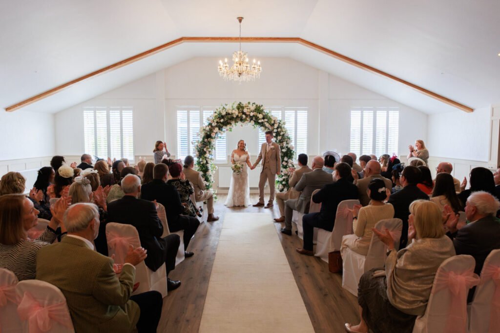 A couple stands hand in hand at the altar during their wedding ceremony, surrounded by guests in a beautifully decorated venue with floral arrangements.