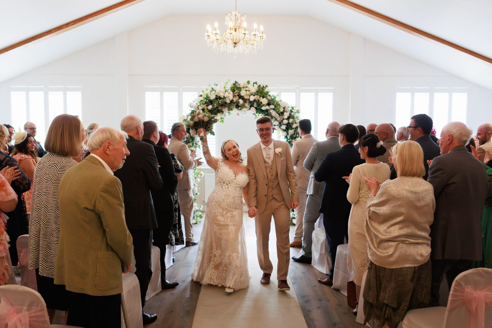 A bride and groom joyfully walking hand in hand down the aisle after their wedding ceremony, surrounded by guests applauding.