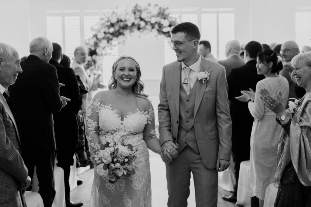 Bride and groom walking hand in hand down the aisle, smiling, surrounded by applauding guests in a floral adorned venue.