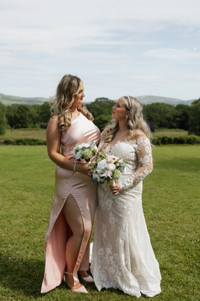 Two women stand in a lush green field, one wearing a pink dress and the other in a white lace wedding gown, both holding bouquets of flowers.