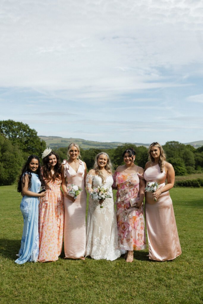 A group of six women standing together outdoors, dressed in elegant dresses, with lush greenery and hills in the background. The bride, in a lace gown, holds a bouquet, while the bridesmaids wear pastel-coloured outfits.
