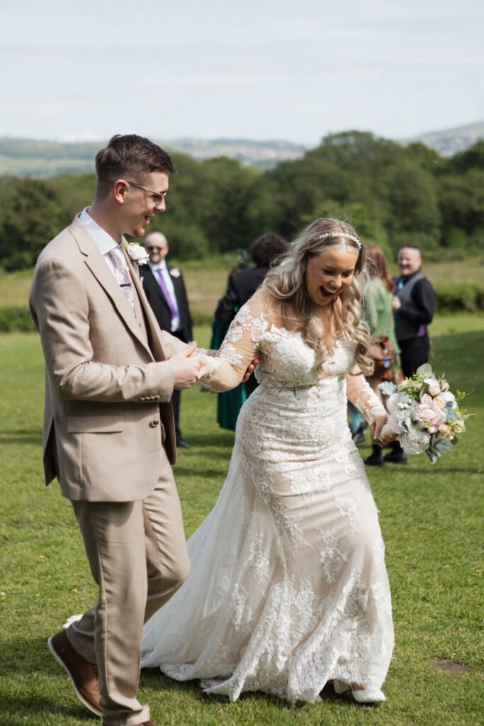 A bride and groom joyfully walking hand-in-hand on a green lawn, surrounded by guests. The bride wears a beautiful lace wedding dress, while the groom is dressed in a light-coloured suit. A sunny day with trees and hills in the background.