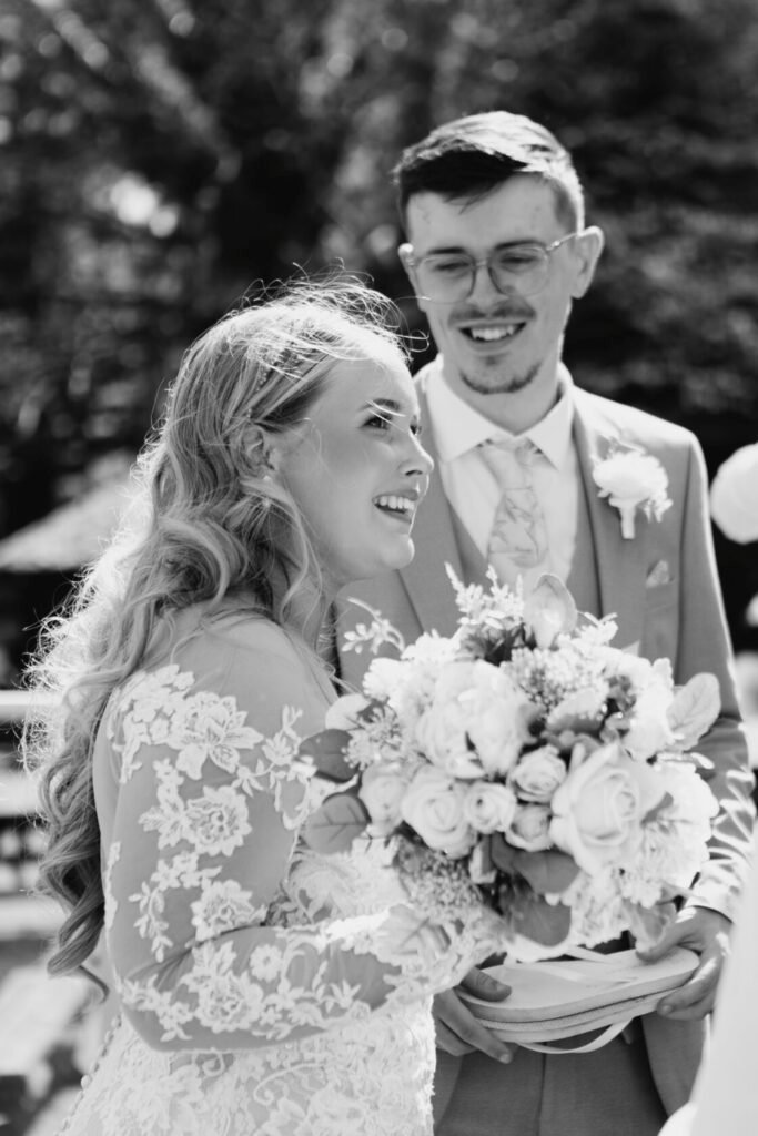 Bride smiling while holding a bouquet, standing next to the groom, in a sunlit outdoor setting.