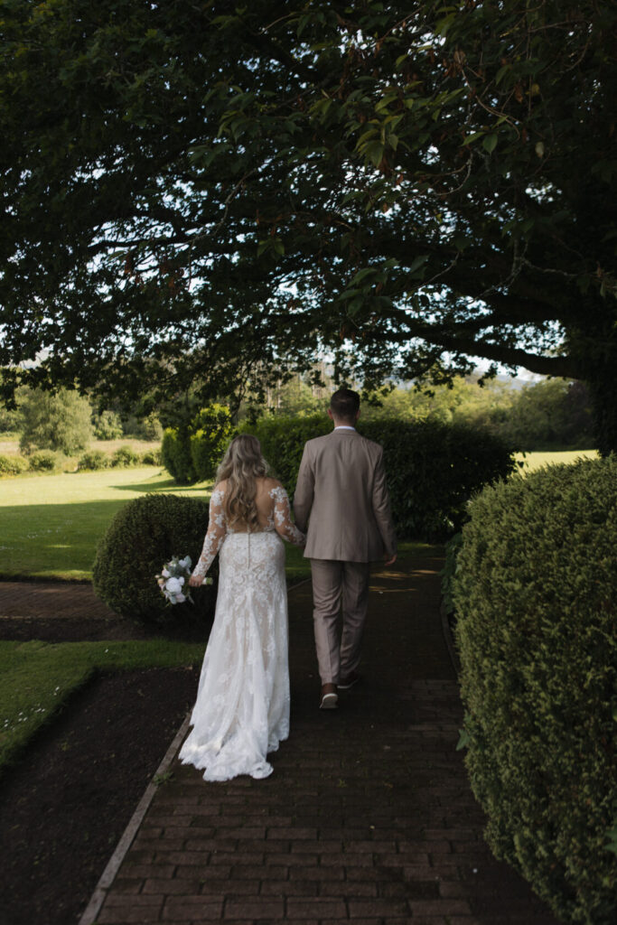 Bride and groom walking hand in hand on a path surrounded by greenery, with the bride wearing a lace wedding gown and holding a bouquet.