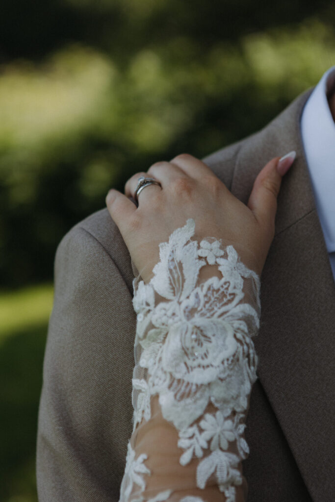 Close-up of a hand with a lace sleeve resting on a brown suit shoulder, showcasing a wedding ring.