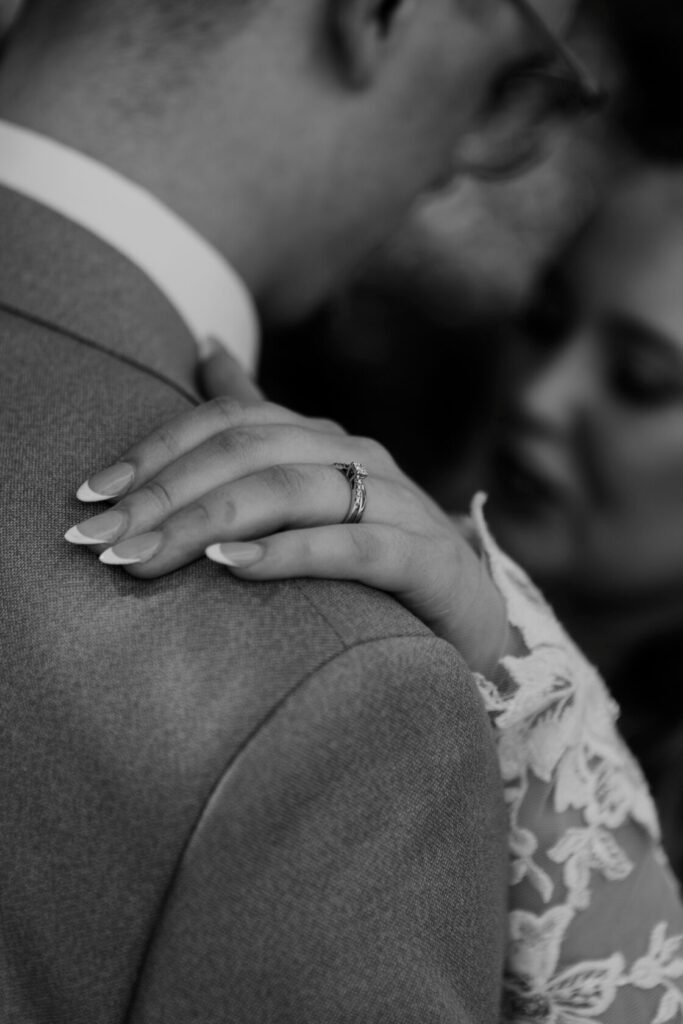 Bride's hand adorned with a wedding ring resting gently on the groom's shoulder, showcasing elegant nails and intricate lace details in the background.