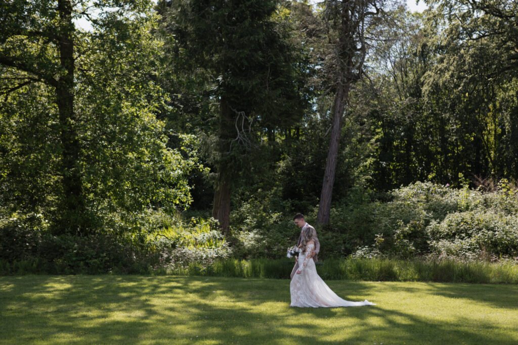 A bride and groom walking hand in hand on a grassy area, surrounded by lush greenery and trees.