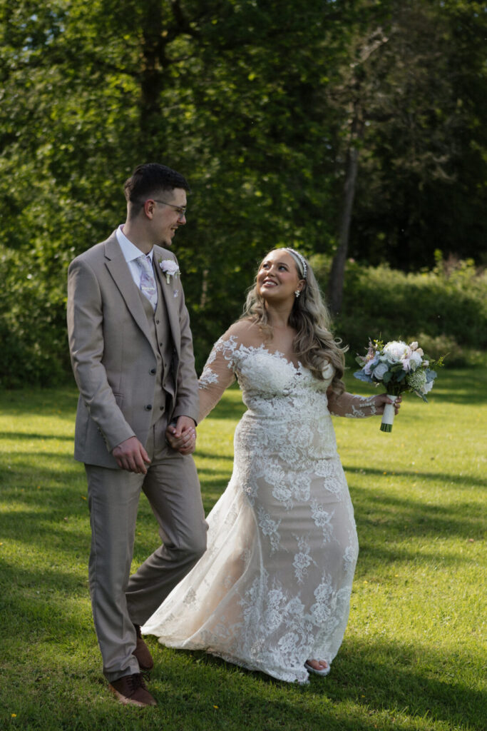 A bride in a lace wedding dress and a groom in a light suit walking hand in hand through a grassy area, surrounded by trees