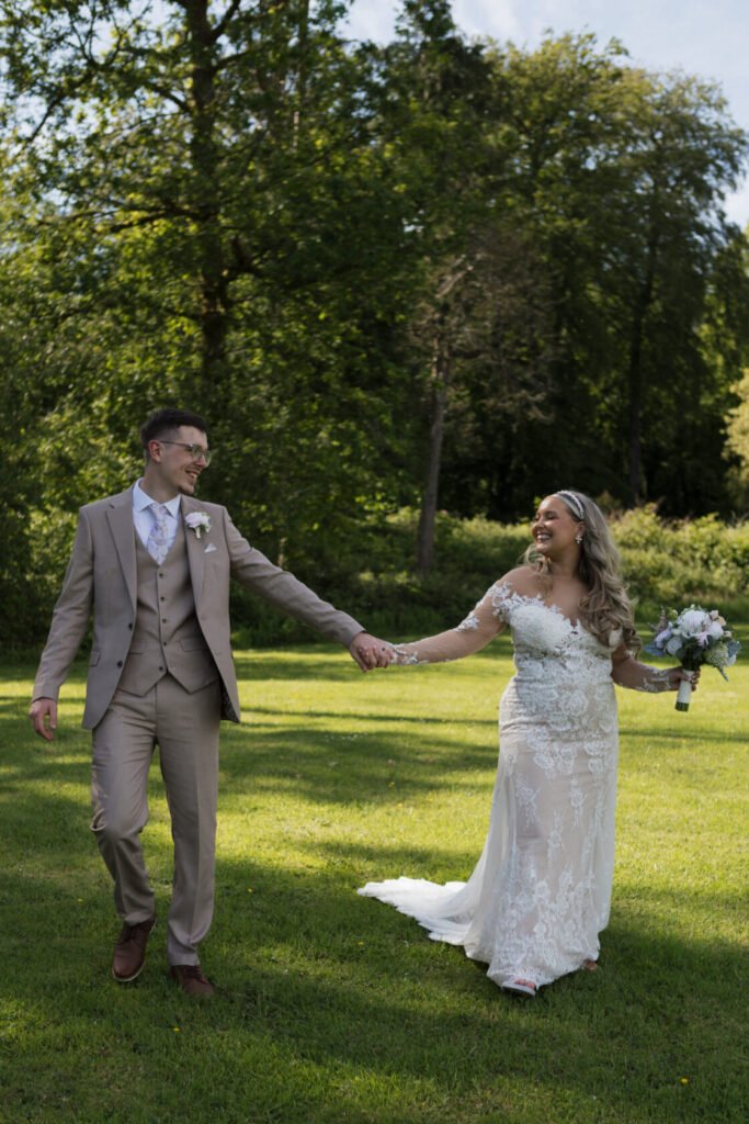 A bride in a lace gown and a groom in a beige suit are walking hand in hand on a lush green lawn, surrounded by trees.