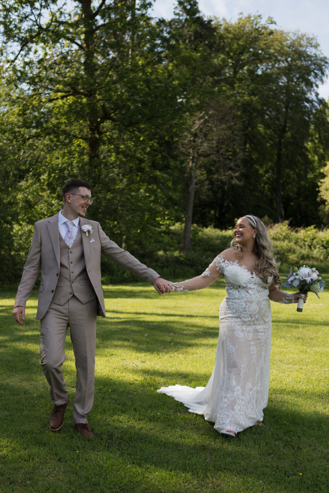 A bride in a lace gown and a groom in a beige suit are walking hand in hand on a lush green lawn, surrounded by trees.