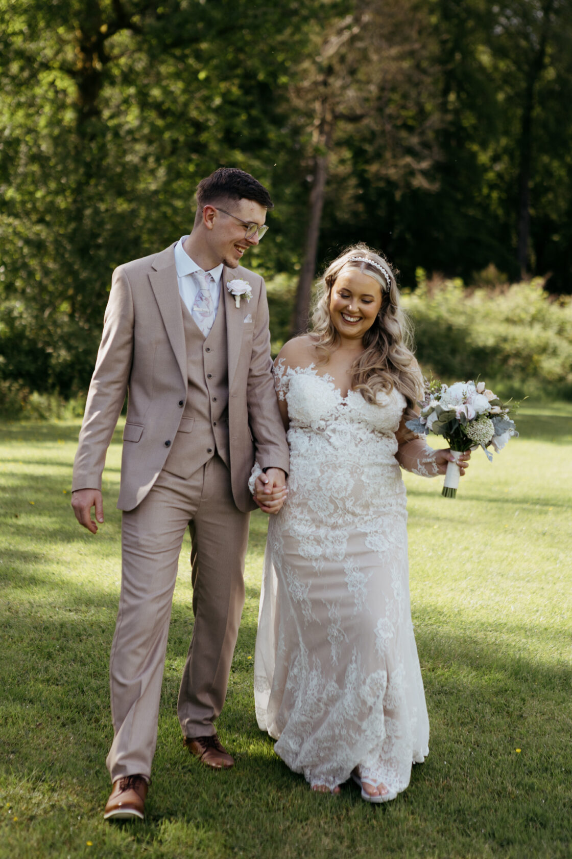 A couple holding hands and walking through a grassy area. The bride wears an elegant lace gown and holds a bouquet, while the groom is dressed in a light beige suit.