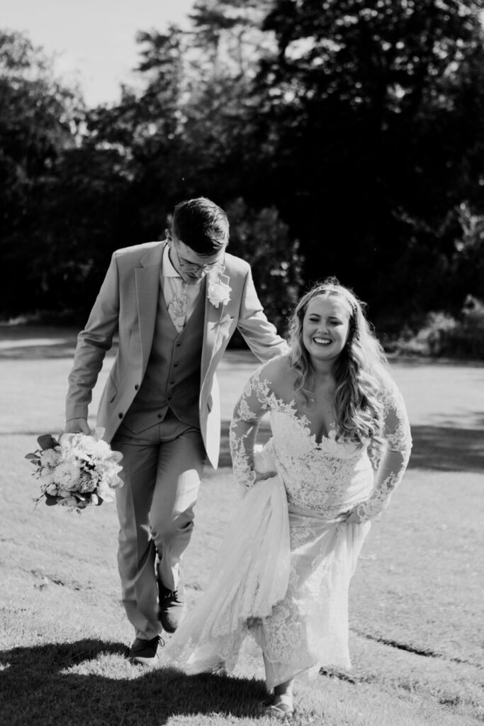 A joyful bride in a lace wedding dress and a groom in a grey suit walking hand in hand on grass, with the bride playfully lifting her dress.