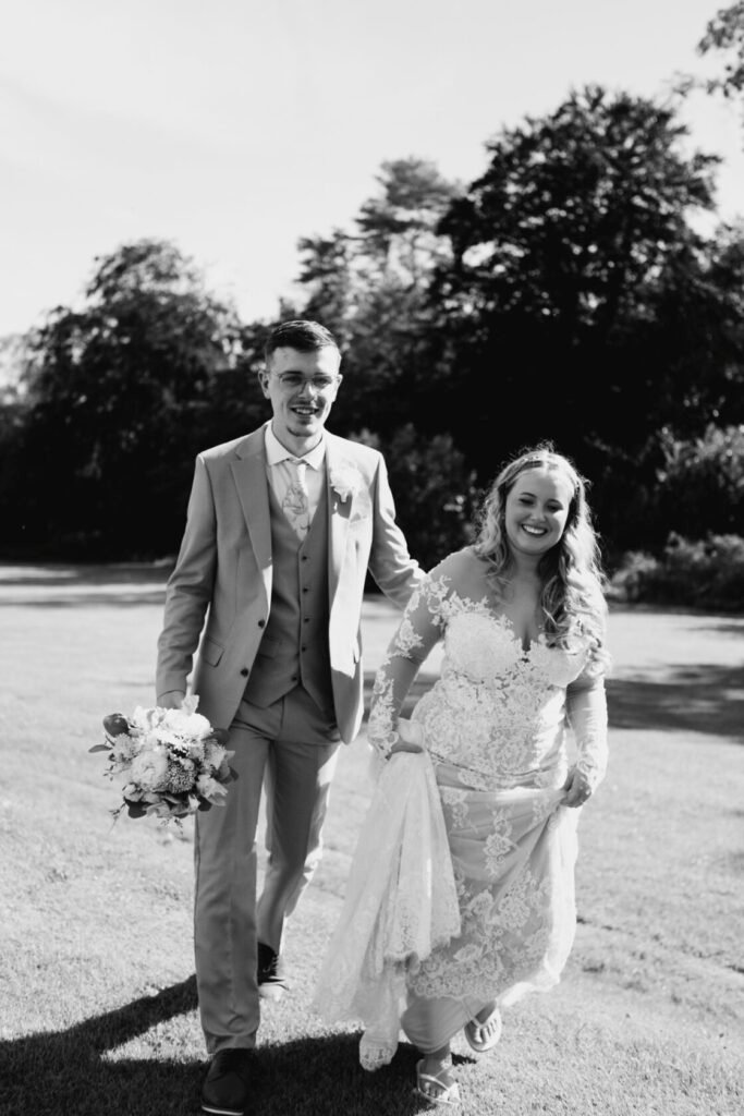 A smiling bride in a lace wedding dress and a groom in a grey suit walking hand in hand, surrounded by greenery.