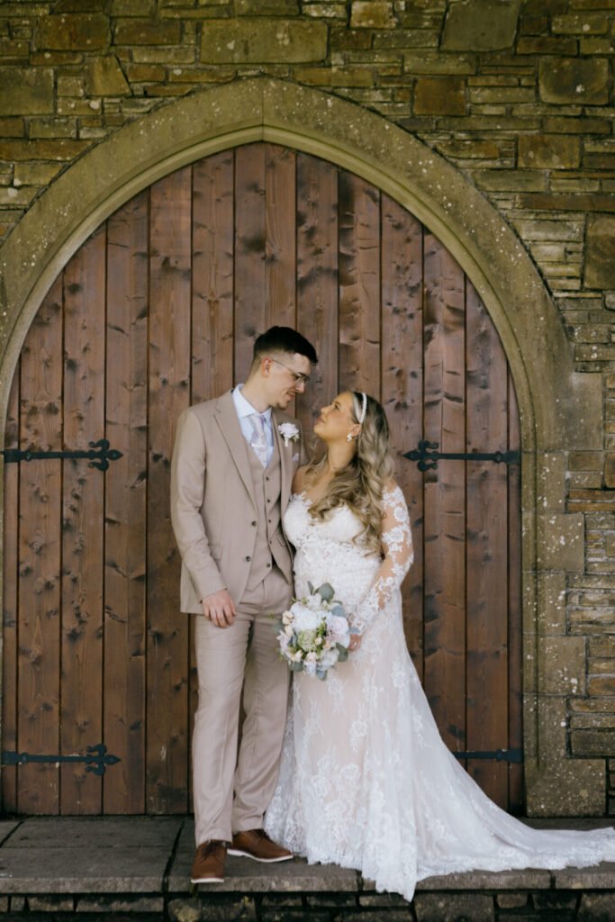A newlywed couple stands close together, gazing into each other's eyes. The groom is dressed in a light tan suit, while the bride wears an elegant lace wedding dress and holds a floral bouquet. Behind them is a rustic wooden door.