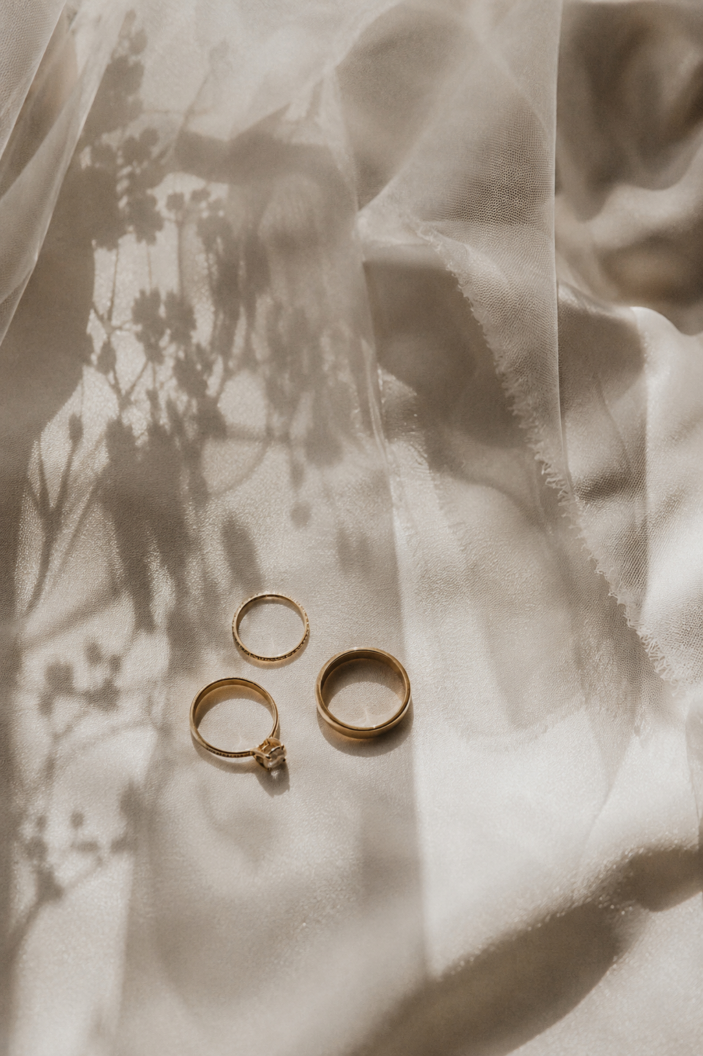 Three gold wedding rings arranged on soft satin fabric, with delicate floral shadows cast in the background. Shot for Lydia Turner.