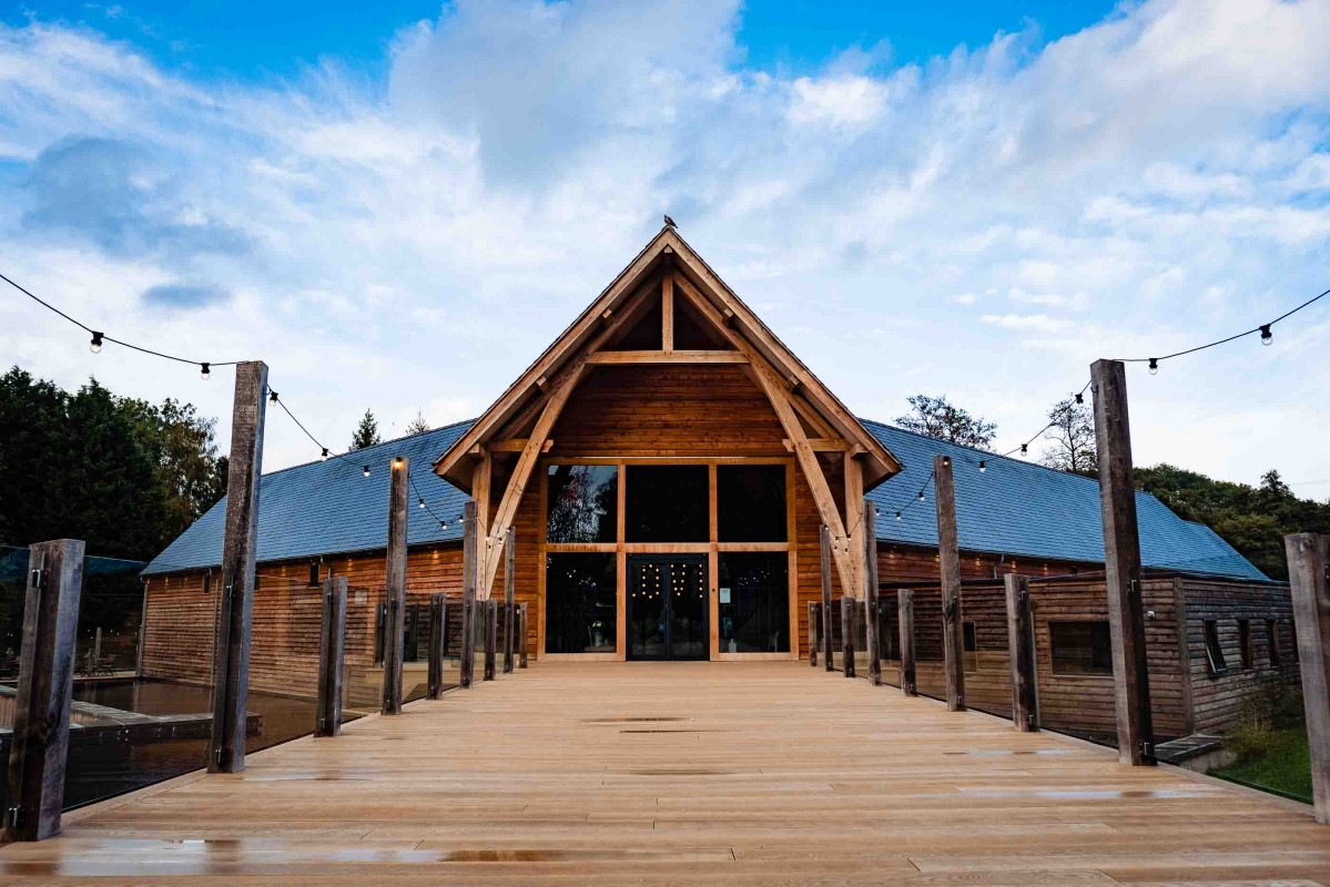 A wooden pavilion with a triangular roof and a large entrance, flanked by a wooden walkway and surrounded by trees under a blue sky.