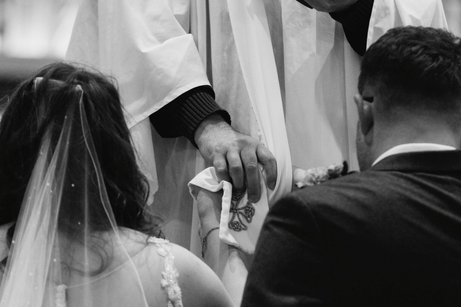 Black and White Wedding Ceremony Moment Bride and groom participating in a wedding ceremony, focusing on a priest's hands holding ceremonial cloth while the couple kneels.