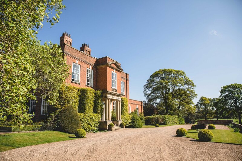 A grand Georgian mansion featuring red brick architecture, surrounded by manicured gardens and trees under a clear blue sky.