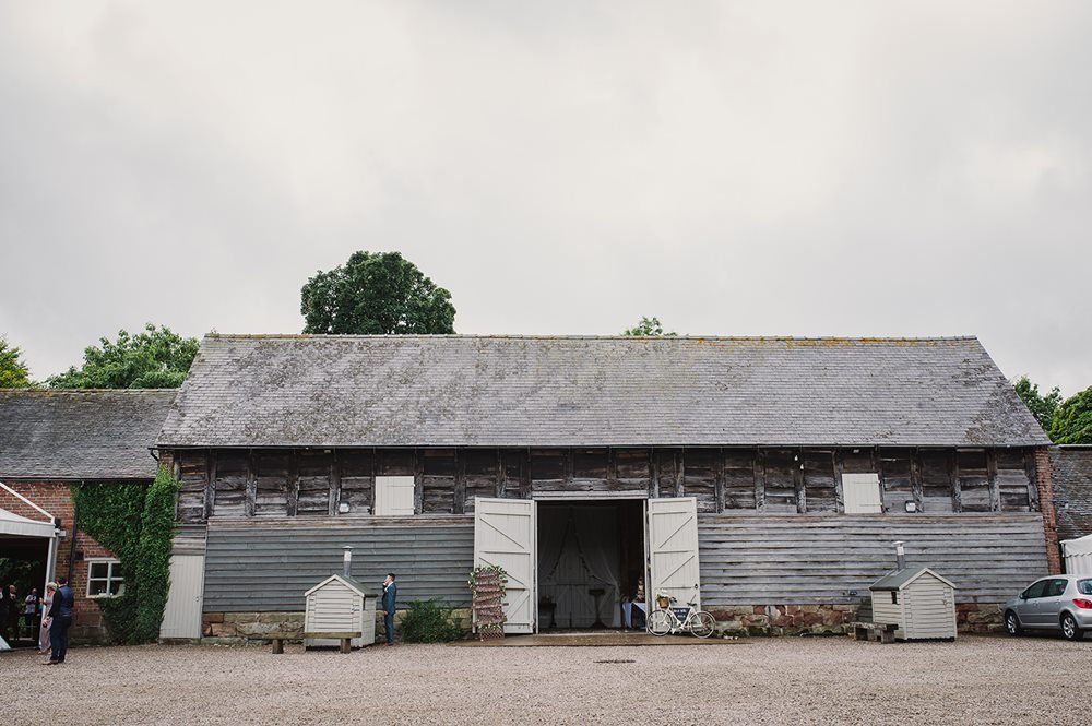 A rustic wooden barn with open doors, surrounded by greenery and small structures, set against a cloudy sky.