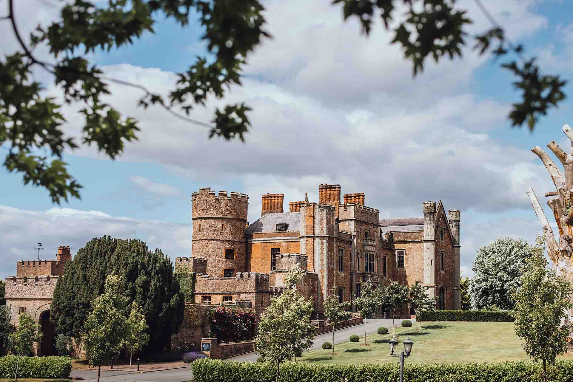 A picturesque castle featuring a mix of stone and brick architecture, surrounded by trees and landscaped gardens under a cloudy sky.
