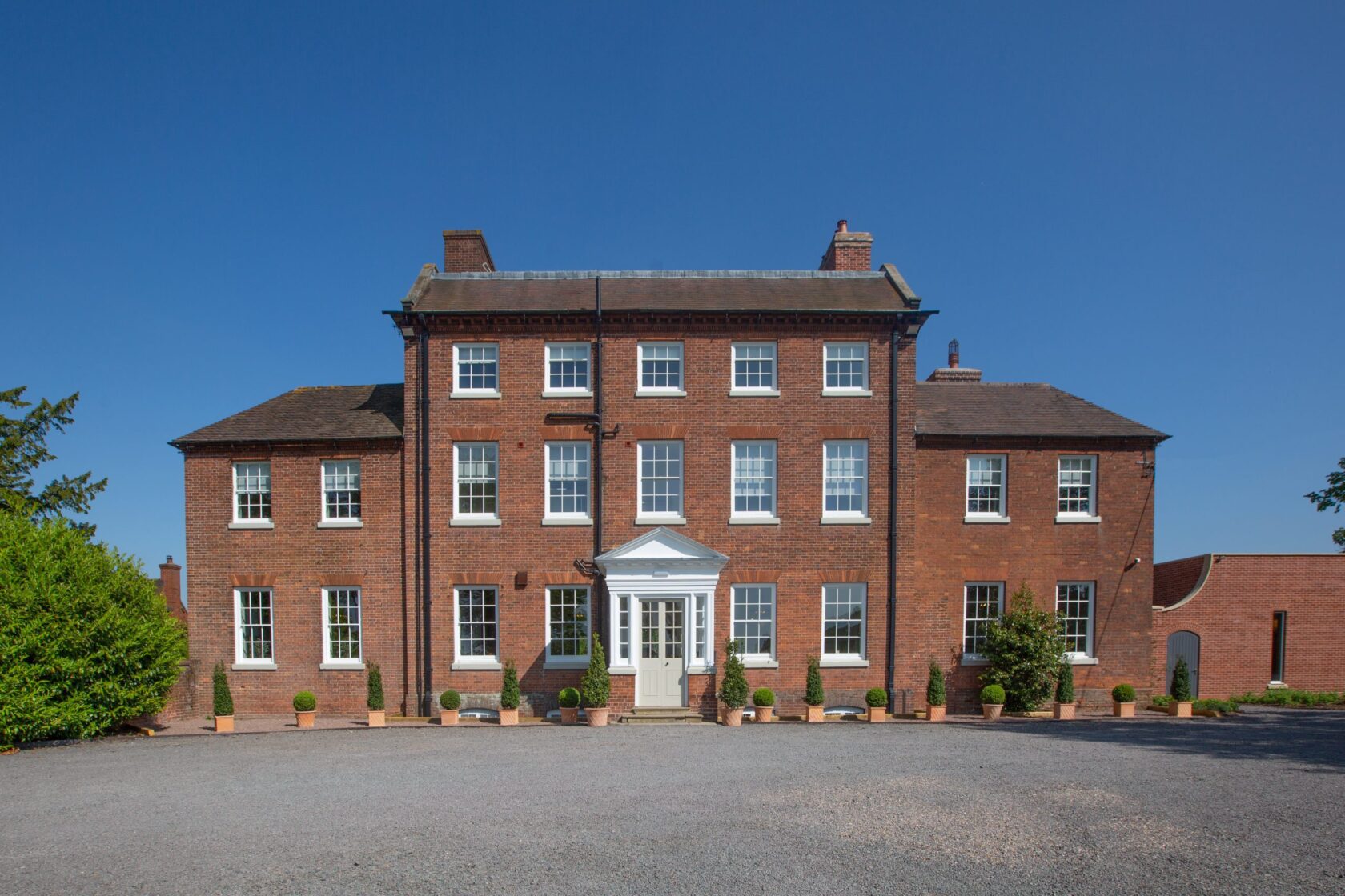 A large, classic red brick country house with white sash windows and a grand entrance, surrounded by greenery and potted plants.