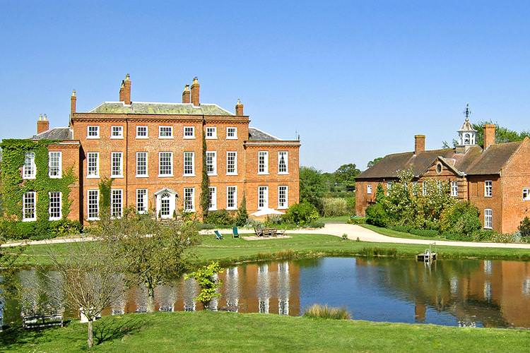 A large red brick manor house with white windows, surrounded by lush greenery and a tranquil pond in the foreground.