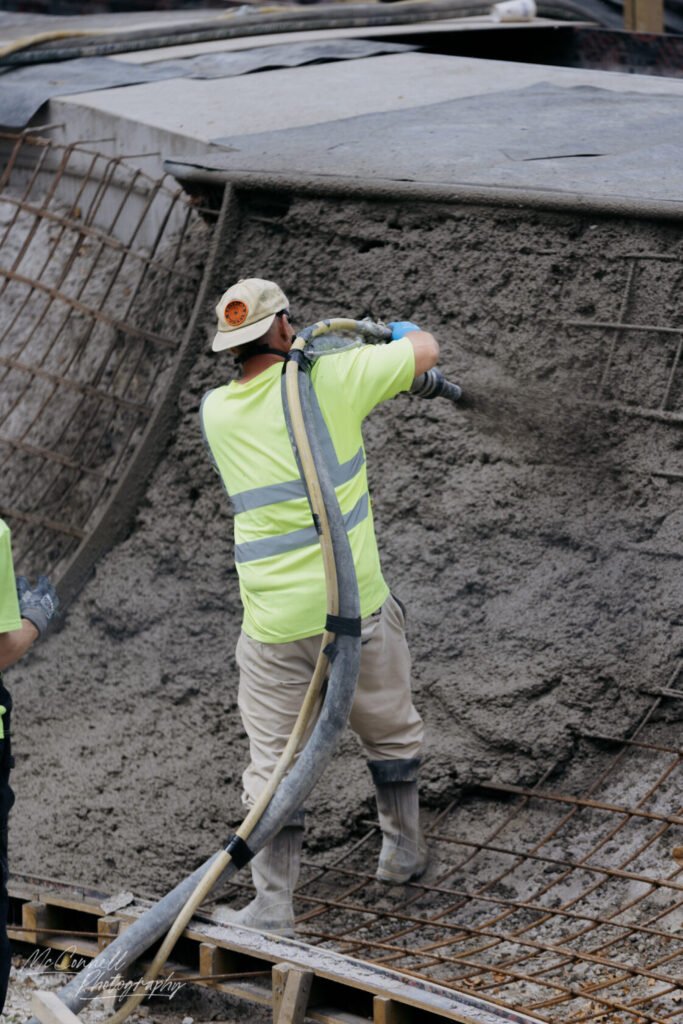 A construction worker in a bright yellow vest and cap pours concrete using a hose at a construction site, with reinforcement bars visible in the background.