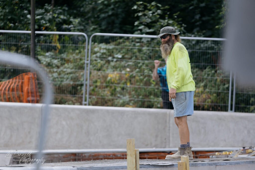 A construction worker with a long beard, wearing a bright yellow shirt and a hat, stands on a construction site, overseeing the work with a fence and greenery in the background.