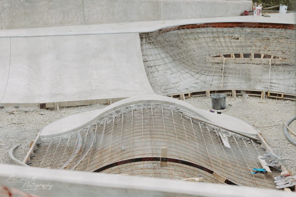 A partially completed skatepark bowl featuring curving concrete and exposed rebar, with construction tools and materials visible on the ground