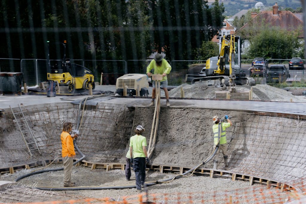 Four construction workers pouring concrete into a curved structure at a construction site, surrounded by machinery and building materials.
