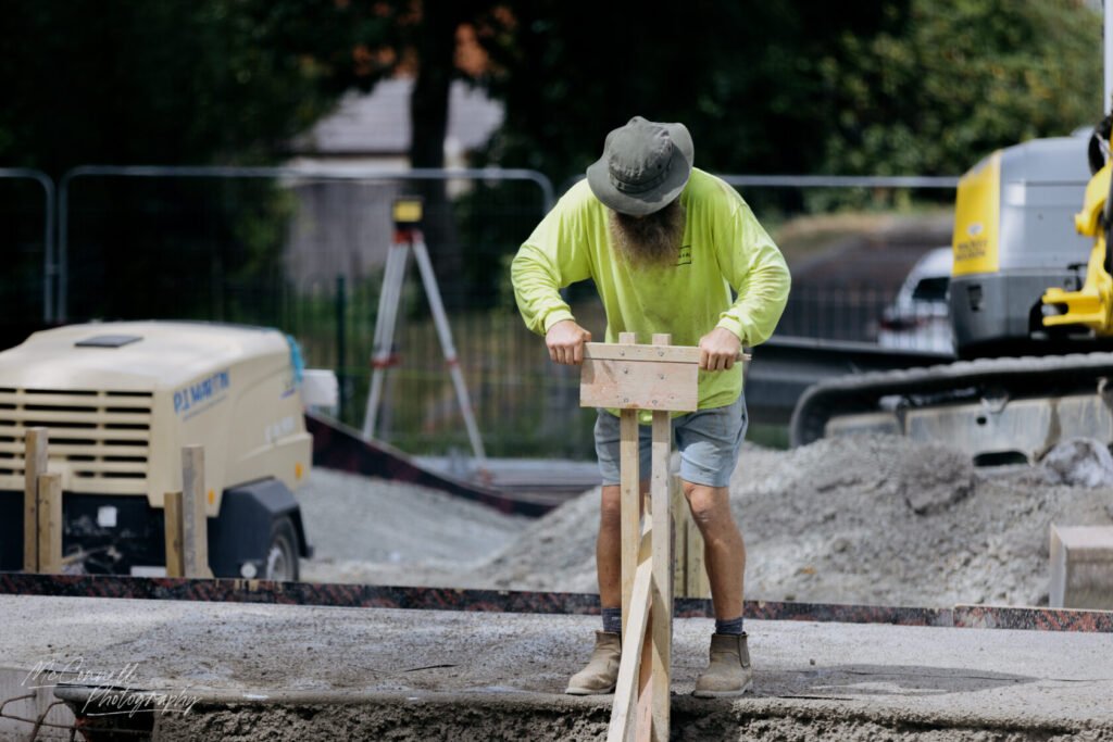 A construction worker with a beard, wearing a bright yellow long-sleeve shirt and shorts, is adjusting wooden formwork on a construction site.