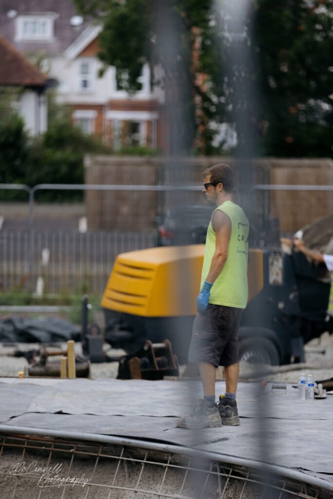 A construction worker wearing a bright yellow vest and gloves stands on a construction site, with machinery and tools in the background, fenced off by barriers.