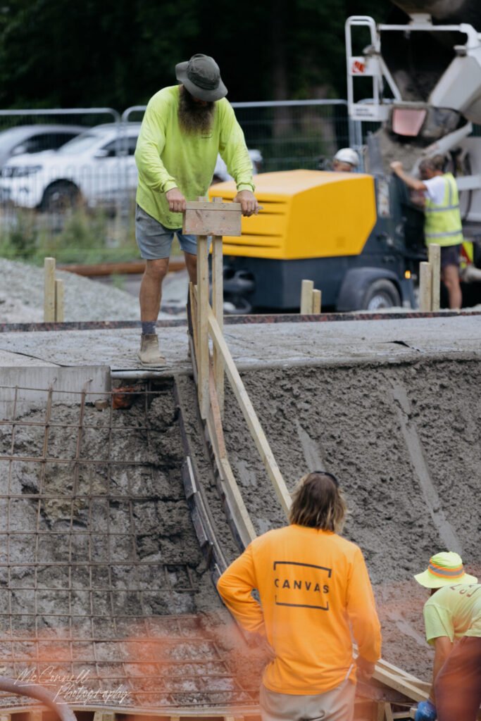 A construction worker in a lime green shirt and hat is levelling concrete using a wooden tool, while others assist in the background amidst construction machinery.
