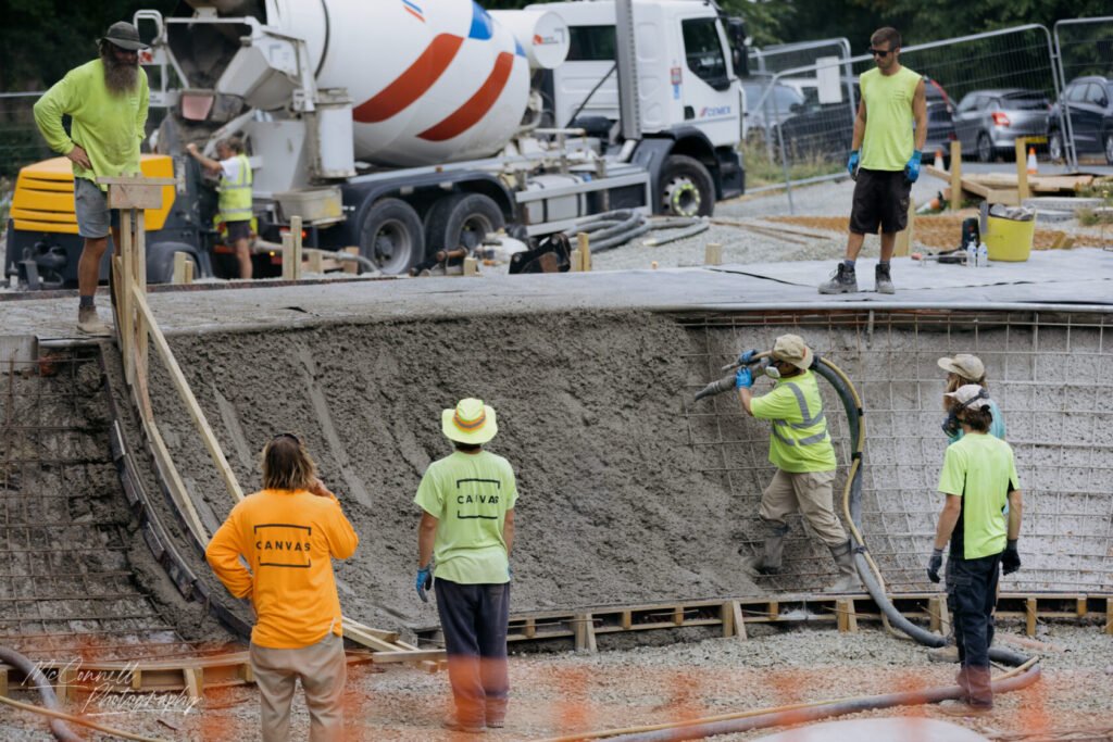 A group of construction workers in brightly coloured safety gear pouring concrete on a building site, with a cement mixer in the background and wooden forms in place.