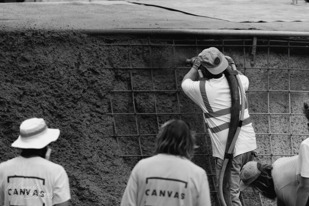 A construction worker using a spraying tool to apply a material on a reinforced surface. Several other workers are visible in the background, all wearing safety gear.