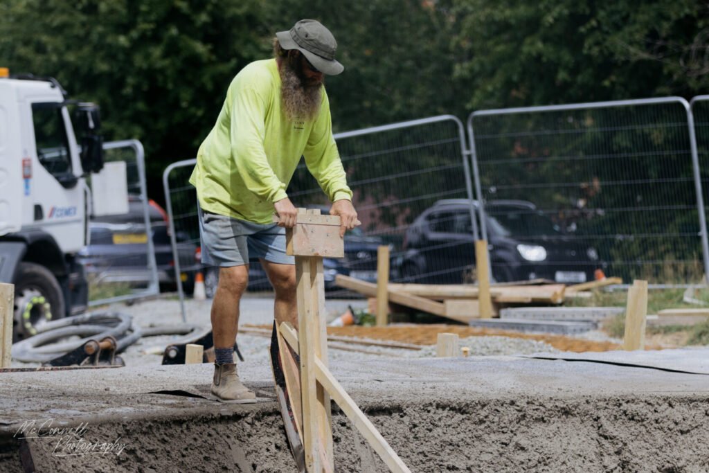 A construction worker in a bright yellow long-sleeve shirt and shorts is adjusting wooden forms for concrete while standing on a construction site with vehicles in the background.