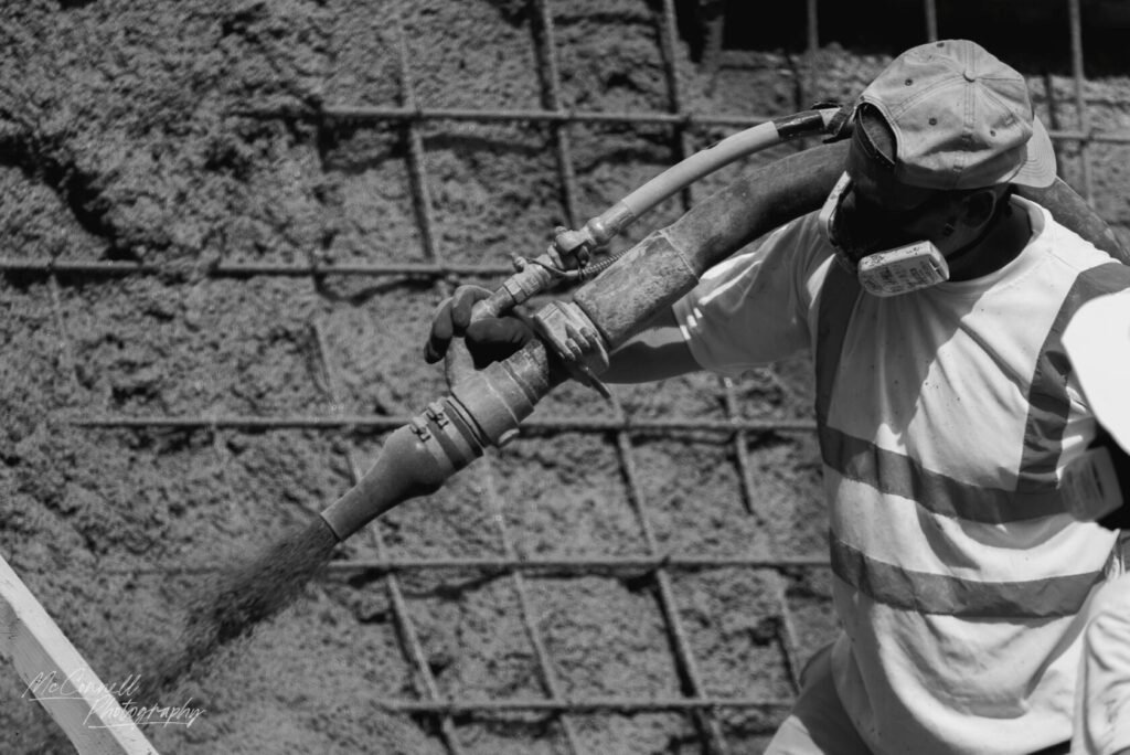 A construction worker operating a concrete sprayer, wearing a mask and cap, amidst a backdrop of textured concrete.