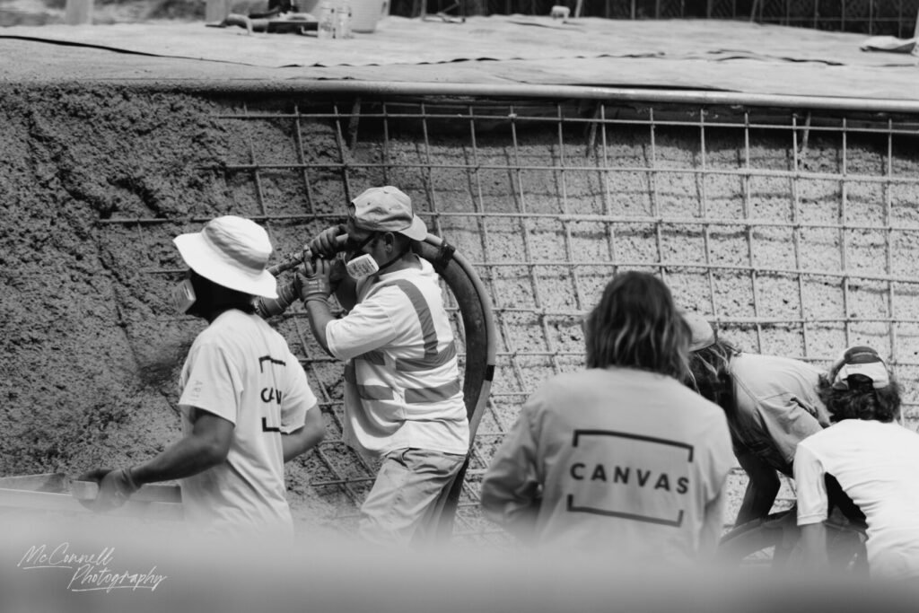 A group of construction workers is using machinery to apply cement to a wall, with one worker holding a hose and others preparing materials.