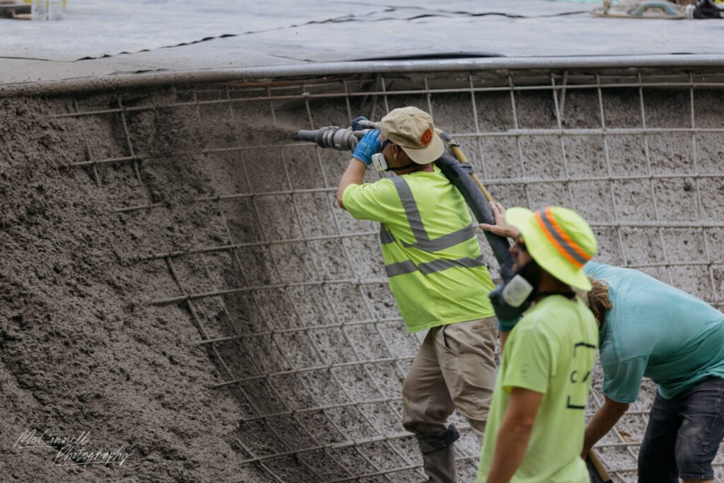 Two construction workers in bright yellow safety vests pouring concrete into a framework, one using a hose and the other assisting.