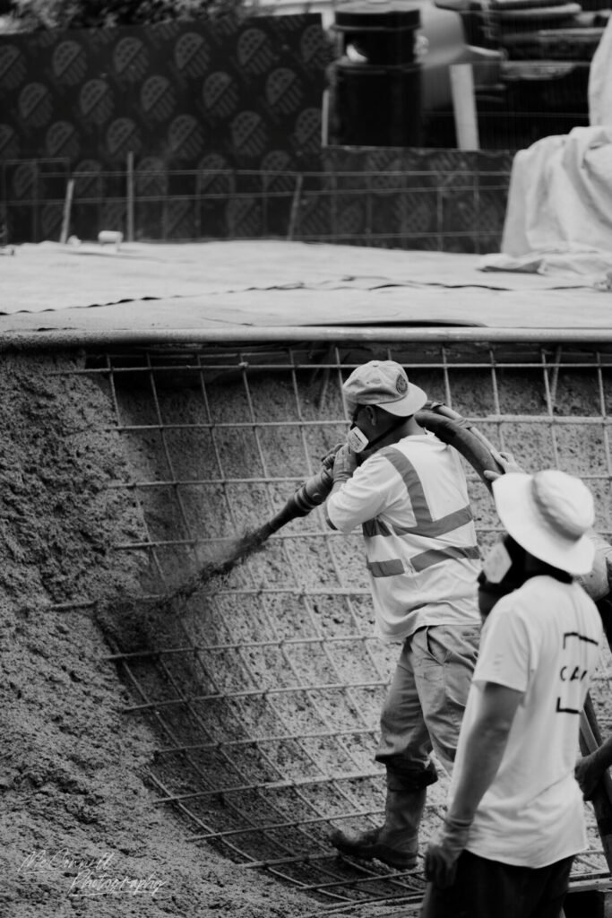 Two construction workers applying concrete to a rebar structure, one using a hose and the other observing.
