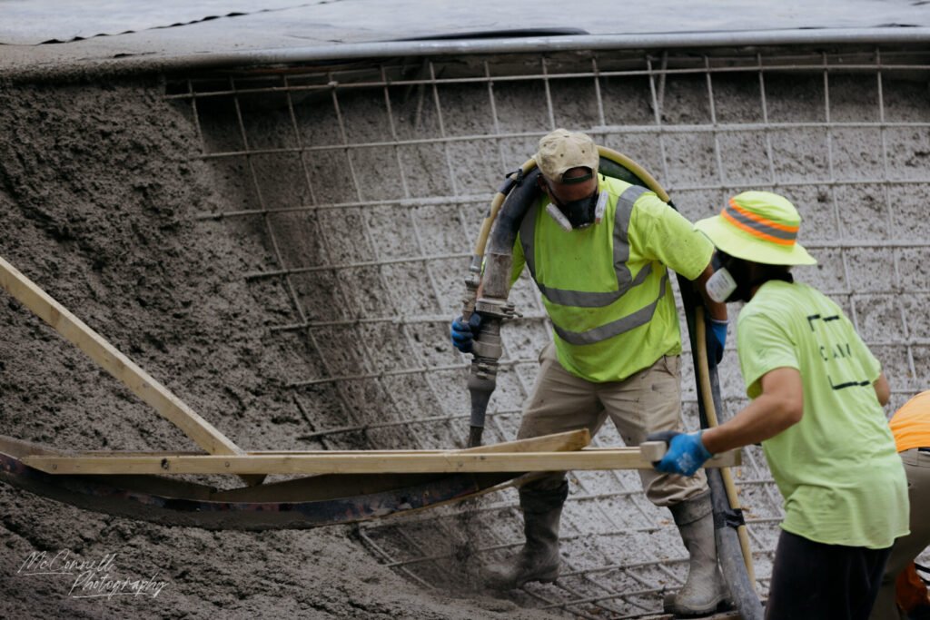 Two construction workers in high-visibility shirts pouring concrete using a hose and a wooden board in a construction site.