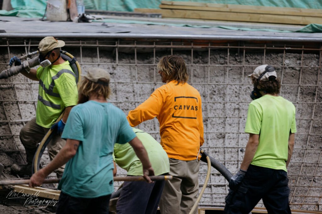 Group of construction workers in safety gear working on a building site, one person operating a concrete hose while others assist.