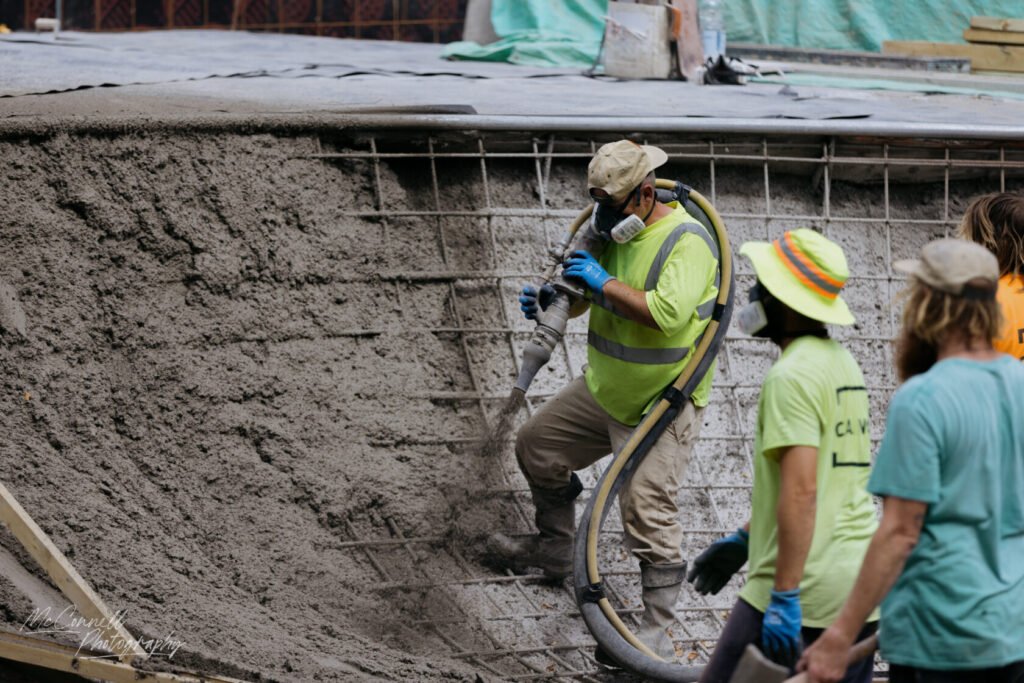 Group of construction workers pouring concrete into a framework. One worker is using a hose to distribute the concrete, while others are assisting and wearing safety gear.
