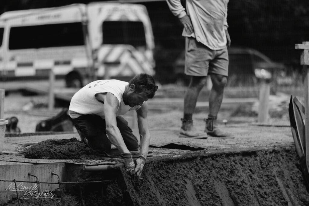 A construction worker kneeling on a concrete slab, handling wet cement, with another worker standing in the background.