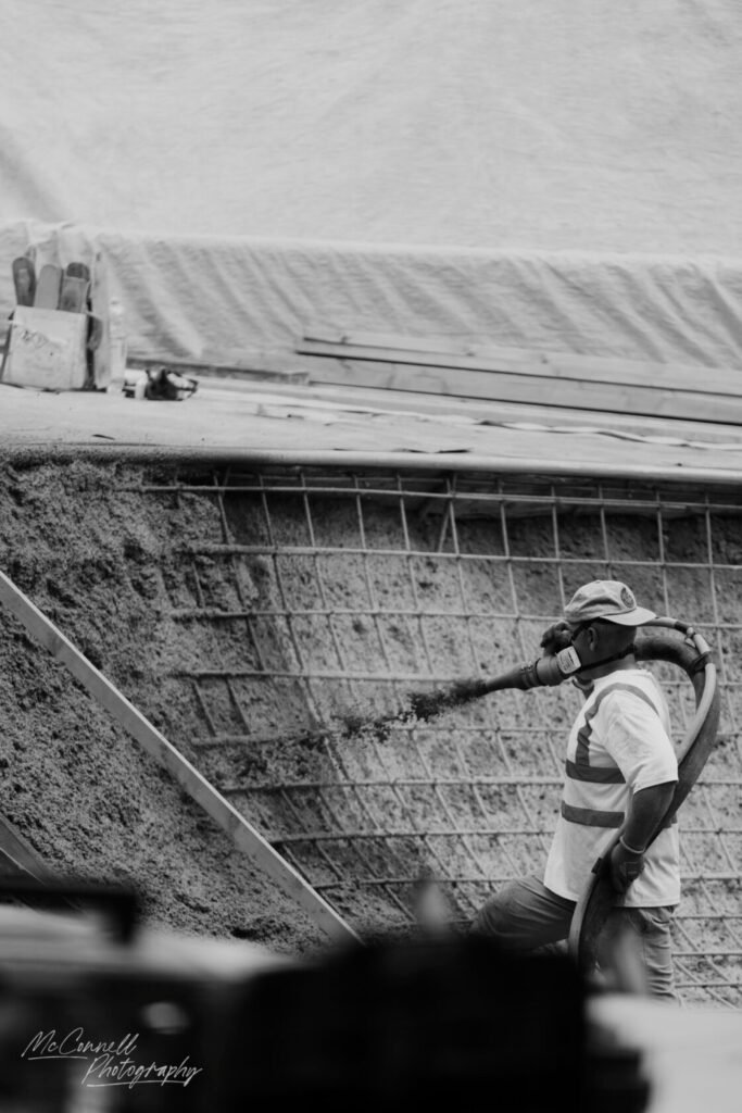 A construction worker operating a hose to pour material onto a building site. The worker is wearing a cap and a reflective vest.
