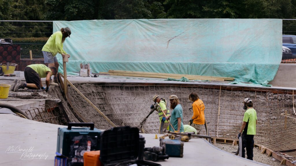 Group of construction workers pouring concrete and working on a pool structure with tools and materials scattered around.
