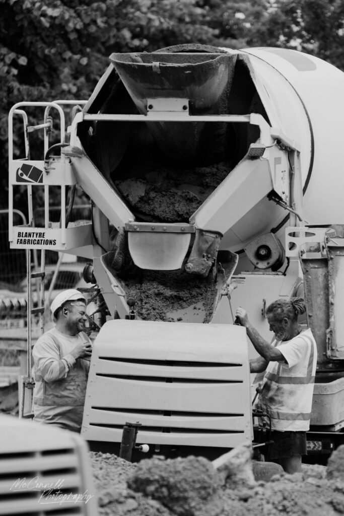 Two construction workers engaged in operating a concrete mixer, one smiling while the other is working on the machinery. The scene is in black and white, featuring details of the mixer and surrounding materials.