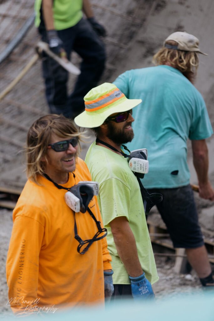 Two construction workers wearing bright clothing and safety gear, smiling while on the job site. One worker is in an orange long-sleeve shirt and the other in a yellow shirt with a wide-brimmed hat.