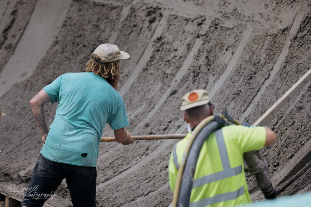 Two construction workers are pouring concrete into a curved wall, one is stirring with a pole and the other is directing the concrete flow through a hose.