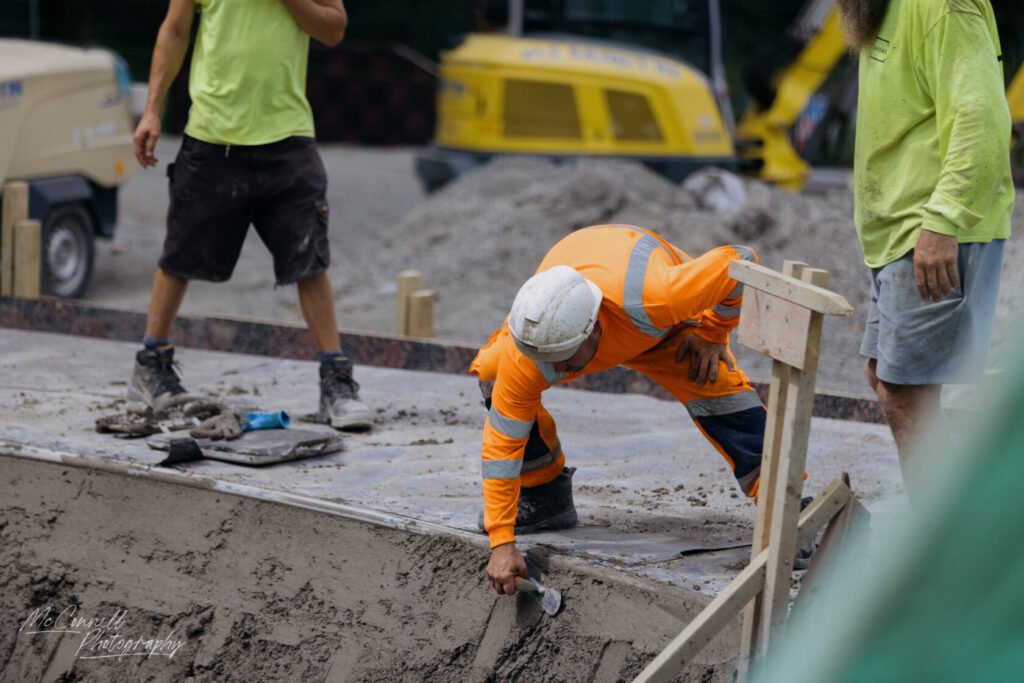 Two construction workers at a building site, one in an orange safety suit kneeling and smoothing wet concrete, and another in a green shirt observing.